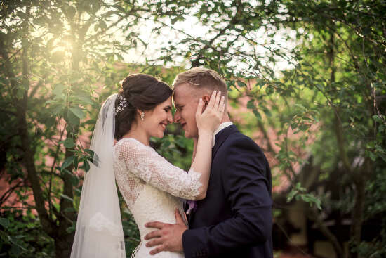 A newlywed couple outside in front of trees, about to kiss.