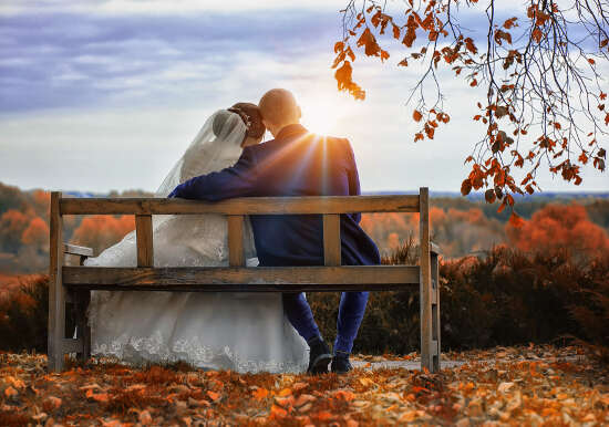 A newlywed couple, from behind, cuddling on a park bench looking toward the horizon, colorful fall leaves on the ground.